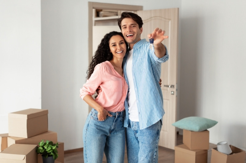 A smiling couple stands close together in a room with moving boxes, with the man pointing playfully towards the camera. The room is bright and has an open door and a plant in the background.