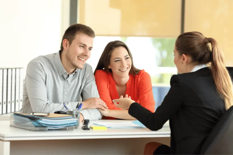 A smiling couple sits at a desk across from a professional woman in an office, having a friendly discussion. The couple appears attentive and engaged, with documents and folders on the desk.